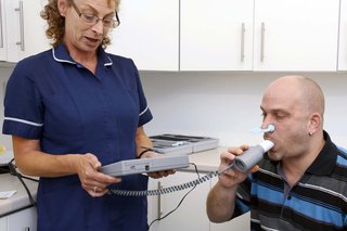A person having a spirometry test. They're seated and have a clip on their nose. They're breathing into a tube attached to a device being held by a healthcare professional.